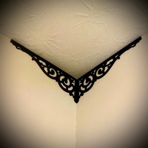 May include: Black metal corner shelf bracket with a decorative, ornate design. The bracket is mounted in the corner of a room, where the wall meets the ceiling.