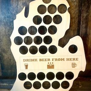 May include: A light-colored wooden wall display shaped like the state of Michigan, featuring numerous circular cutouts for bottle caps. The board includes the engraved text "DRINK BEER FROM HERE" along with small icons of a glass, a crate, and a beer mug.