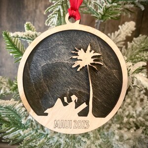 May include: A wooden Christmas ornament with a silhouette of a palm tree and a person sitting on a beach. The ornament is painted black and has the text "MAUI 2023" engraved on the bottom.