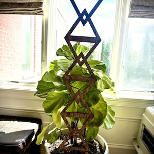 May include: A geometric wooden plant trellis in a white pot with a large green plant. The trellis, made of dark brown wood, features diamond and triangle shapes. The plant pot is near a window. The plant is a Fiddle Leaf Fig.