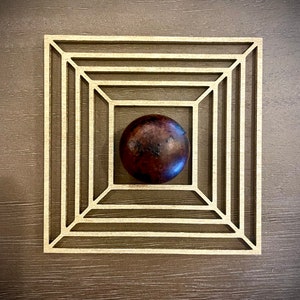 May include: A dark brown sphere sits in the center of a wooden square frame with multiple concentric squares.