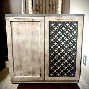 May include: A white cabinet with two doors. The left door is solid wood, while the right door has a black metal lattice design. The cabinet has a gray countertop and a silver faucet.