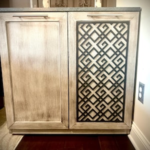 May include: A white wooden cabinet with two doors. The right door has a black geometric design cutout. The cabinet has silver handles.