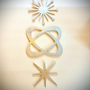 Könnte beinhalten: Drei silberfarbene Metall-Wandkunstwerke. Das oberste Stück ist ein Sternenmuster, das mittlere eine verschlungene Form und das untere ein Stern. Alle sind an einer hellen Wand befestigt.