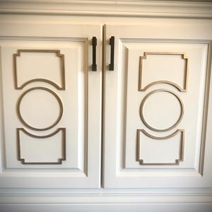 May include: Two white cabinet doors with gold trim and black rectangular handles. The doors have a decorative design with a circle and a square shape.