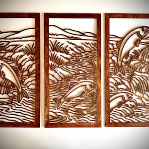May include: A set of three wooden wall art panels. Each panel features a laser-cut design of a fish leaping out of a river, with a backdrop of mountains and vegetation. The panels are framed in dark brown wood.
