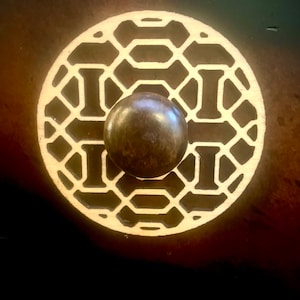 Puede incluir: Un pomo de cajón redondo de madera con un diseño geométrico. El pomo está pintado de oro y tiene un centro de metal marrón.