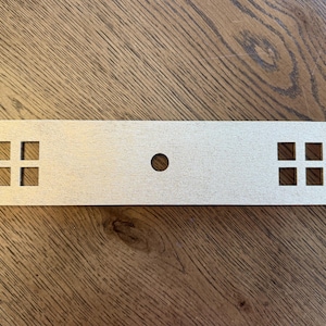 May include: A rectangular, light-colored wooden plaque featuring a smooth, flat surface. The piece includes a single circular hole centered in the middle and two square-shaped cutouts on each end, each containing four smaller square openings.