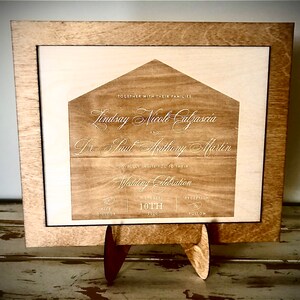 May include: A wooden wedding invitation with a house-shaped design. The invitation is engraved with the names "Lindsay Nicole Calzascia" and "Dr. Paul Anthony Martin" and the date "October 10th".