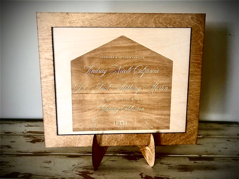 May include: A wooden wedding invitation with a house-shaped design. The invitation is engraved with the names "Lindsay Nicole Calzascia" and "Dr. Paul Anthony Martin" and the date "October 10th".