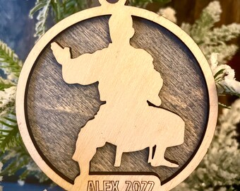 May include: A wooden Christmas ornament with a silhouette of a person in a karate pose. The ornament is brown with a wood grain pattern and has the name "ALEX 2022" engraved on the bottom.