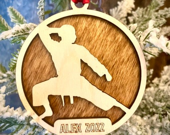 May include: A wooden Christmas bauble featuring a silhouette of a person in a karate pose. The bauble is brown with a wood grain texture and has the text "ALEX 2022" engraved on the bottom.