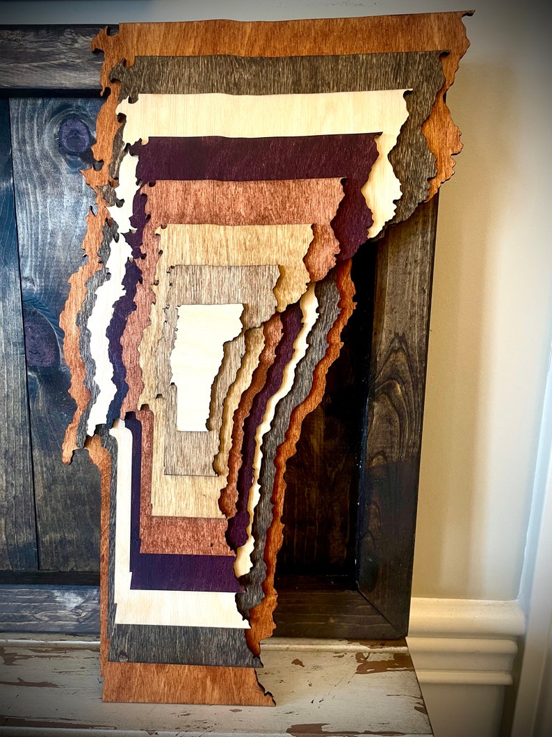 Puede incluir: Una pieza de arte de pared de madera en capas con forma del estado de Vermont. La madera est&aacute; te&ntilde;ida en varios tonos de marr&oacute;n, incluyendo marr&oacute;n oscuro, marr&oacute;n claro y marr&oacute;n rojizo. La pieza est&aacute; montada en un marco de madera marr&oacute;n oscuro.