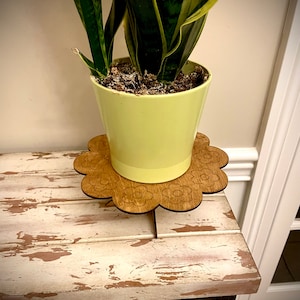 May include: A light green potted plant with long, striped leaves sits on a wooden plant stand with a flower design. The stand has a scalloped edge and is made of light brown wood.