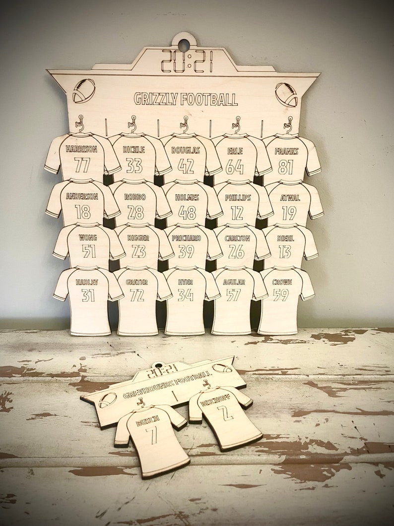 May include: A wooden football jersey holder with the text "Grizzly Football" and "20:21" at the top. The holder has 18 jersey slots with numbers and names printed on each slot. The bottom of the holder has a smaller wooden sign with the text "20:21 Grizzly Football" and two jersey slots with numbers.