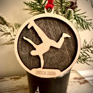 May include: A wooden Christmas ornament featuring a silhouette of a person doing a handstand. The ornament is brown with a dark wood grain and has the text "ERICA 2024" engraved on the bottom.