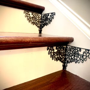 Puede incluir: Primer plano de una escalera con escalones y contrahuellas de madera oscura. Soportes decorativos de metal negro con un diseño en espiral están fijados a los escalones. Los soportes añaden un elemento decorativo a la escalera.