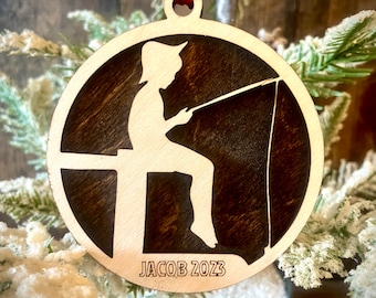 May include: A round wooden ornament with a silhouette of a person fishing. The ornament is brown with a dark brown stain. The text "JACOB 2023" is engraved on the bottom of the ornament.