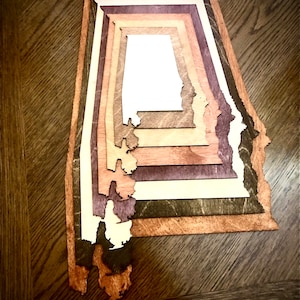 以下が含まれることがあります： アラバマ州の州の形をした木製の切り抜きで、茶色、ベージュ、紫の異なる色の木を重ねています。切り抜きは濃い茶色の木製の表面にあります。