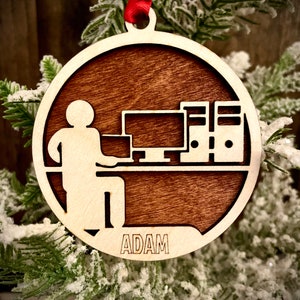 May include: A wooden Christmas bauble featuring a silhouette of a person seated at a desk with a computer, and the name "ADAM" inscribed at the base.