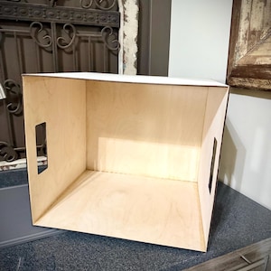 May include: A light-colored, open wooden box with a rectangular shape. The box has two rectangular cutouts on the sides and is placed on a dark countertop. The background includes a decorative metal gate and a distressed wooden frame.