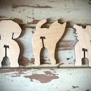 May include: Three wooden cutouts of a dog, cat, and another dog, all facing away from the viewer. The cutouts are in the shape of the animals' rear ends, with the tails facing up. The cutouts are painted in a light wood color.