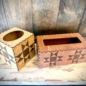 May include: Two wooden tissue box covers. One is square with a circular opening, the other is rectangular with a slot. Both have a light wood finish with dark star-shaped cutouts. The boxes sit on a distressed white surface.