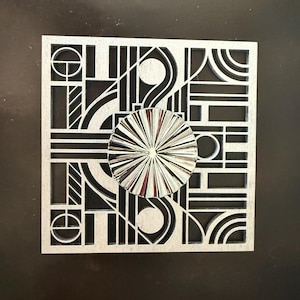 May include: A square silver-toned decorative panel featuring a complex geometric cutout pattern of intersecting lines, circles, and arcs. A circular, metallic, sunburst-textured medallion is centered on the front of the intricate lattice frame.
