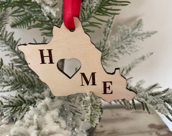 Puede incluir: Un adorno navideño de madera con forma de casa con la palabra "HOME" escrita con un corazón en lugar de la "O". El adorno está colgado de una cinta roja en un árbol de Navidad artificial nevado.