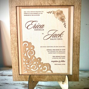 May include: A wooden laser-cut wedding invitation with a floral design and the text "Mrs. Lynda Newman requests the honor of your presence at the marriage of her daughter Erica Danielle to Jack Ralph Tawil Son of Mr. Ralph Tawil and Mrs. Ellen Tawil Sunday, the sixth of June two thousand and twenty-one eleven-thirty in the morning The Crystal Plaza Livingston, New Jersey reception to follow Semi-formal attire"