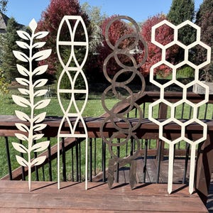 May include: Four wooden plant trellises standing in a row. The first is a leaf pattern, the second features an arched geometric design, the third consists of interlocking dark circles, and the fourth displays a repeating hexagonal honeycomb pattern.