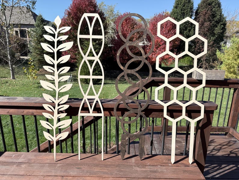 May include: Four wooden plant trellises standing in a row. The first is a leaf pattern, the second features an arched geometric design, the third consists of interlocking dark circles, and the fourth displays a repeating hexagonal honeycomb pattern.