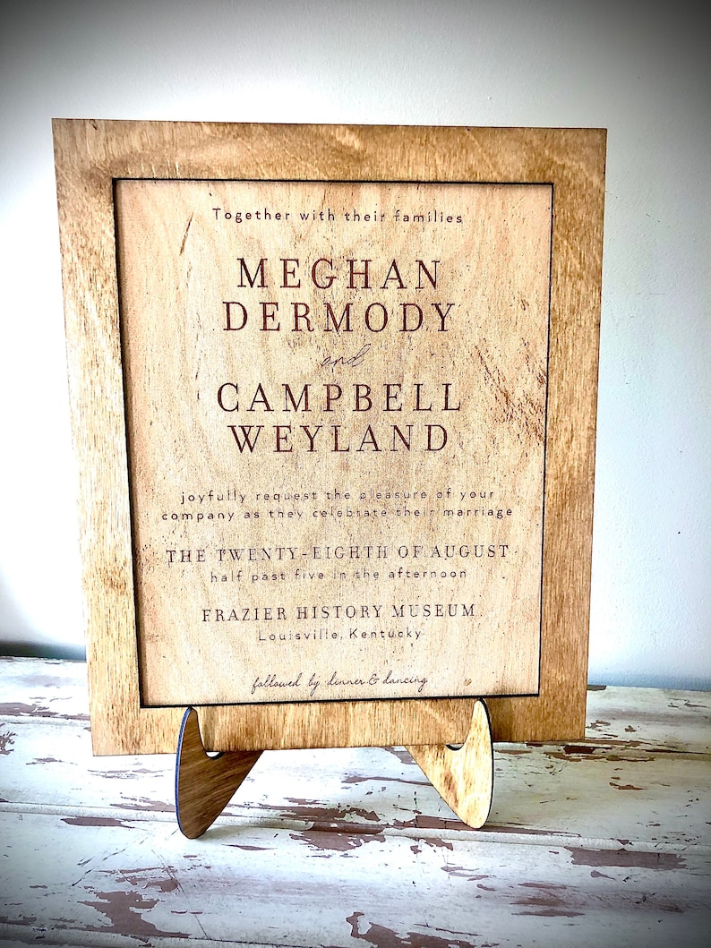 May include: A rustic wooden sign with a cream background and brown text announcing the wedding of Meghan Dermody and Campbell Weyland. The wedding will take place on August 28th at 5:30 PM at the Frazier History Museum in Louisville, Kentucky. The sign is followed by the text "followed by dinner & dancing."