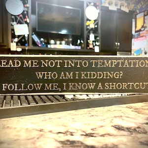 May include: A black wooden sign with gold lettering that reads "Lead me not into temptation... Who am I kidding? Follow me, I know a shortcut". There is a small apple and a stick figure with a trident on the sign.