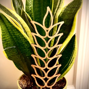 May include: A wooden plant stake shaped like a wheat stalk with a brown color. The stake is in a green pot with a green plant.