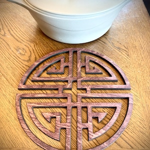 Puede incluir: Un salvamanteles redondo de madera con un diseño geométrico en tono marrón rojizo. Se encuentra sobre una superficie de madera, con una olla de color claro con tapa en el fondo. El salvamanteles está diseñado para proteger las superficies de la cocina caliente.