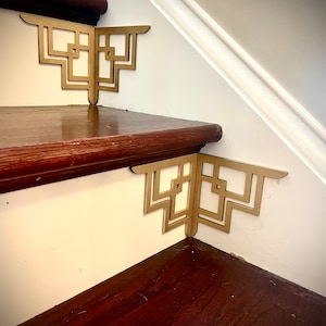 May include: Two gold metal corner brackets with a geometric design are attached to the edge of a wooden staircase. The brackets are in the shape of a right angle and have a repeating pattern of squares and rectangles.