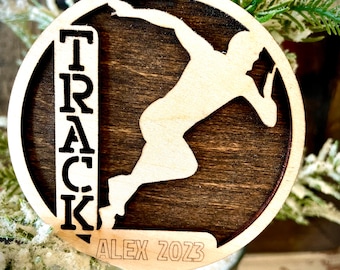Peut inclure: Ornement de Noël en bois avec un thème d'athlétisme. Il présente une silhouette découpée d'un athlète en action, le mot "TRACK" verticalement aligné, le nom "ALEX" et l'année "2023". Un ruban rouge est attaché pour l'accrocher.
