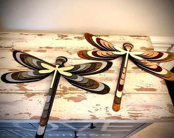 May include: Two wooden dragonfly wall decor pieces with layered wings in shades of brown, gray, and beige. The dragonflies are mounted on a whitewashed wooden surface.