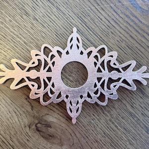 May include: A flat, copper-colored metal plate featuring an intricate, symmetrical, openwork filigree design with a circular cutout in the center. The decorative piece has pointed, leaf-like extensions on both sides and top and bottom.