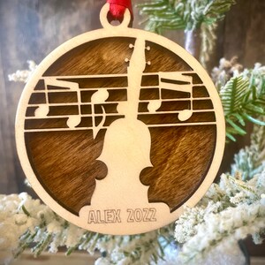 Puede incluir: Un adorno navideño de madera con una silueta de violín y notas musicales. El adorno es marrón y tiene el texto "ALEX 2022" grabado en la parte inferior.
