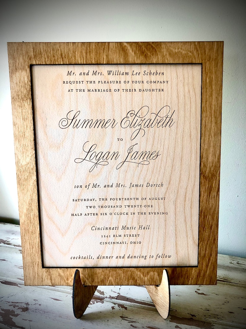 May include: A wooden wedding invitation with a brown frame and a light wood background. The invitation reads "Mr. and Mrs. William Lee Scheben request the pleasure of your company at the marriage of their daughter Summer Elizabeth to Logan James son of Mr. and Mrs. James Dortch Saturday, the fourteenth of August two thousand twenty-one half after six o'clock in the evening Cincinnati Music Hall 1141 BLM Street Cincinnati, Ohio cocktails, dinner and dancing to follow."