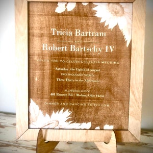 May include: A wooden frame with a light brown background and white text. The text reads "Tricia Bartram and Robert Bartschy IV Invite you to celebrate their wedding Saturday, the Eighth of August Two Thousand Twenty Three Thirty in the Afternoon Allardale Lodge 401 Remsen Rd. Medina, Ohio 44256 Dinner and Dancing to Follow".