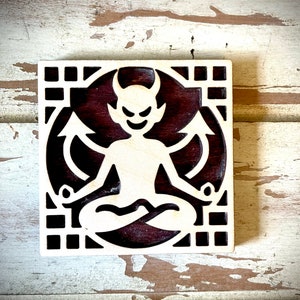 May include: A wooden square with a decorative design featuring a devil figure with horns and a tail, sitting in a meditative pose. The devil is carved in a light wood color, while the background is a dark wood color.