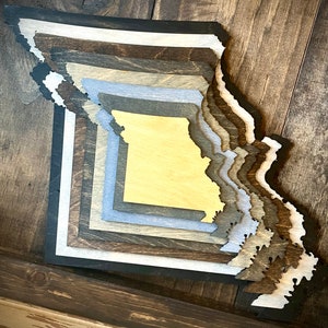 May include: A layered wooden cutout of the state of Missouri. The cutout is made of different shades of wood, including brown, gray, and white. The cutout is on a wooden background.