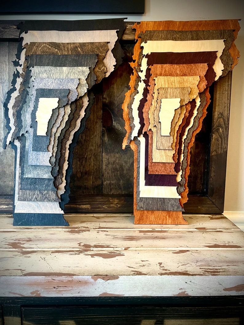 Puede incluir: Dos contornos de estado de madera, uno en tonos marrones y el otro en tonos naranjas, est&aacute;n apilados uno encima del otro. Los contornos se muestran sobre una superficie de madera r&uacute;stica.