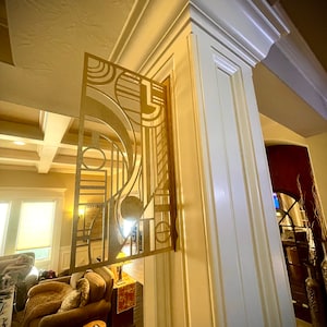 Op de afbeelding: Een gouden metalen wandkunstwerk met een geometrisch ontwerp. De kunst is gemonteerd op een witte muur en is gedeeltelijk zichtbaar door een deuropening.