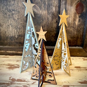 Puede incluir: Tres árboles de Navidad de madera con intrincados diseños recortados. Los árboles son en tonos de marrón, beige y blanco. Cada árbol tiene una estrella en la parte superior.