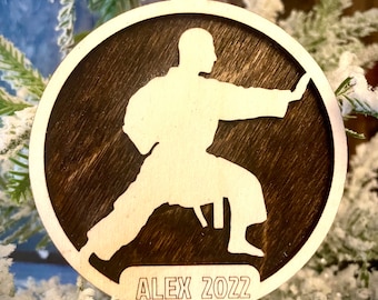 May include: A round wooden ornament with a silhouette of a person in a karate pose. The ornament is brown with a wood grain pattern and has the text "ALEX 2022" inscribed on the bottom.