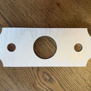 May include: A rectangular light-coloured wooden plate with scalloped edges, featuring one large circular hole in the centre and two smaller circular holes positioned symmetrically on either side of the central opening.